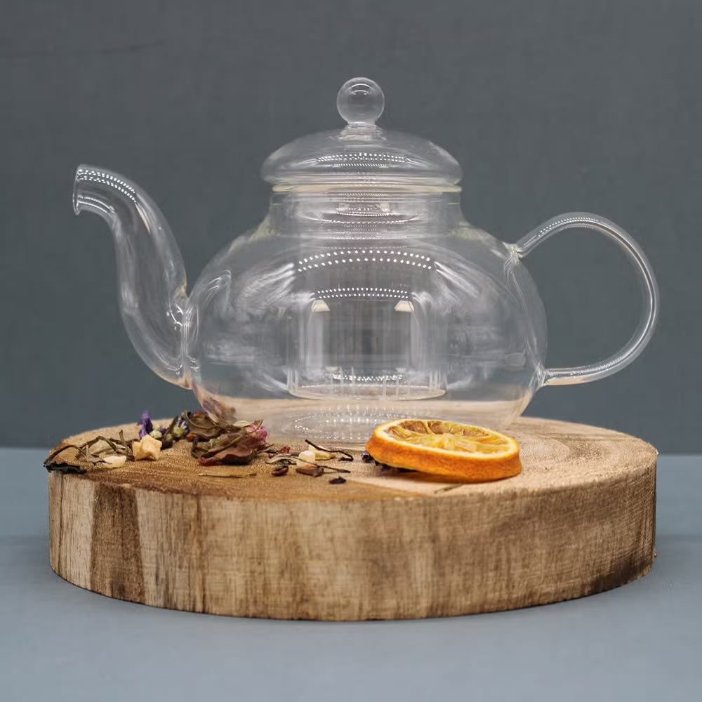 Clear glass teapot on a wooden stand with dried oranges and herbs against a gray background