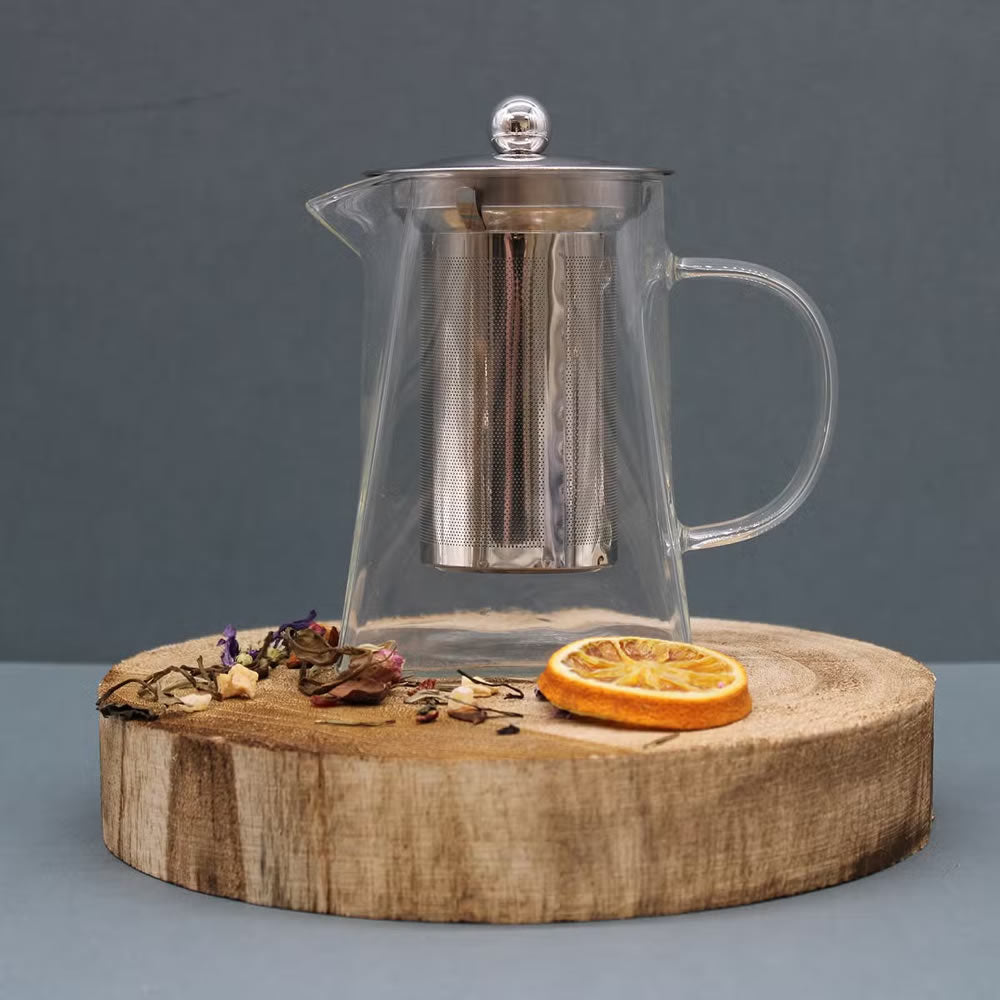 Clear glass teapot with metal infuser on a wooden coaster with dried flowers and an orange slice.