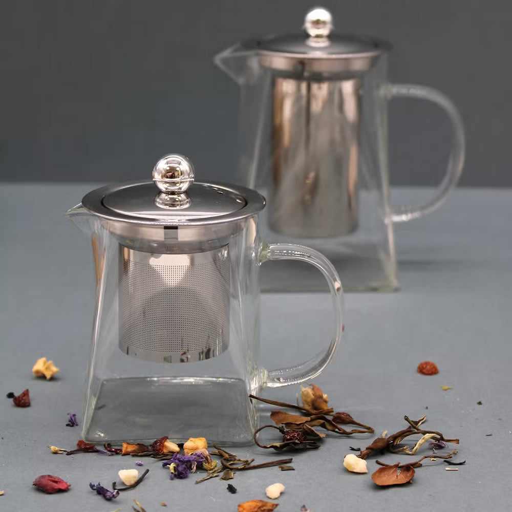 Clear glass teapot with metal lid and mesh filter on a gray surface with dried herbs.