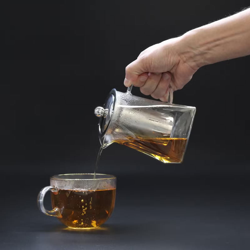 Hand pouring tea from a glass teapot into a glass cup on a black background