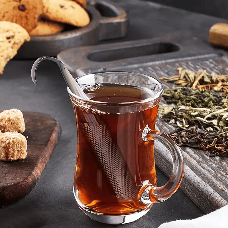 Glass mug of tea with a spoon, surrounded by dried tea leaves and cookies on a wooden surface.